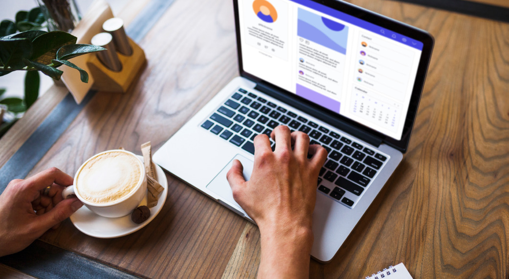 The picture shows a computer on a table. Next to it on the left side is a coffee cup and two hands.